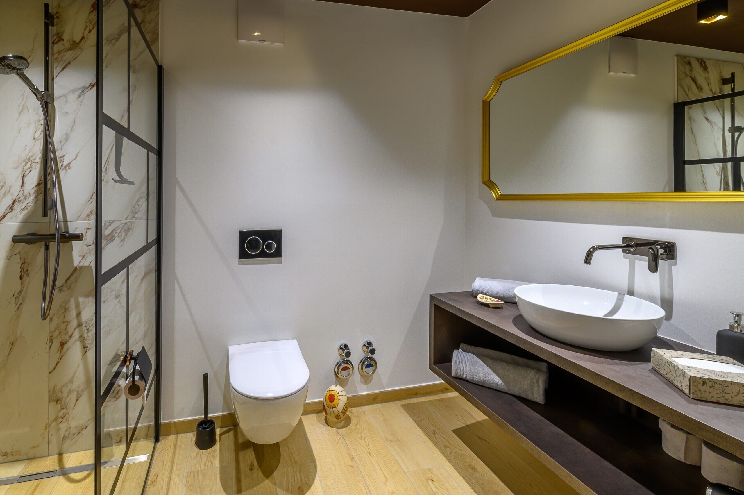 A modern bathroom with a glass shower enclosure, a white toilet, a large vessel sink, and a mirror with a gold frame.