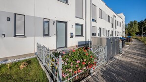 Een modern gebouw met een witte gevel, nummer 57, met een hekwerkentree, een kleine tuin met roze bloemen en een verhard pad.