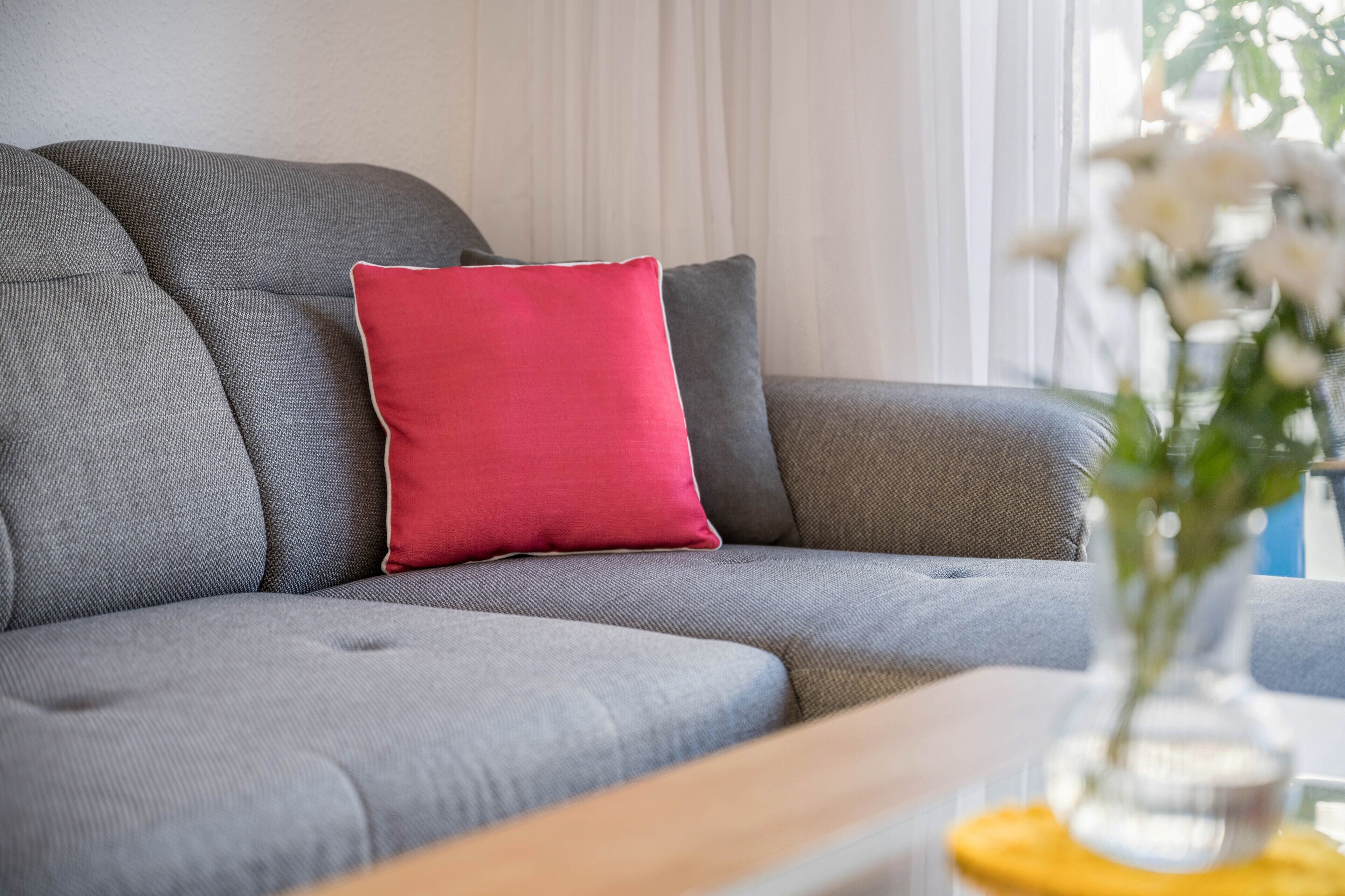 A grey sofa with a red pillow, a wooden side table, and a vase with white flowers.