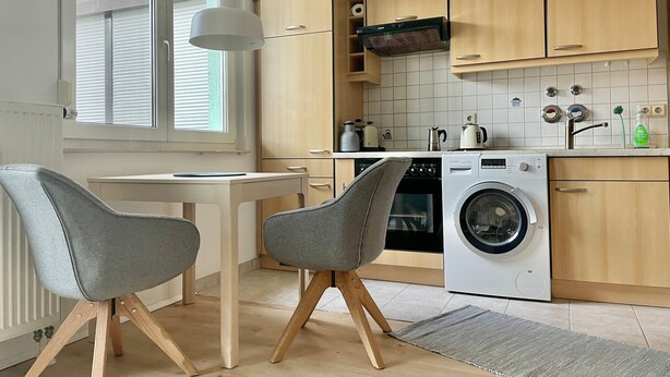 Moderni keittiö, jossa on puiset kaapit, pesukone, kahvinkeitin ja pöytä tuoleineen.
