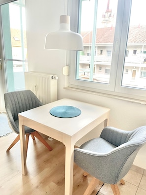 Une pièce avec une table blanche, deux chaises grises, un set de table bleu, une lampe blanche et une fenêtre donnant sur des immeubles.