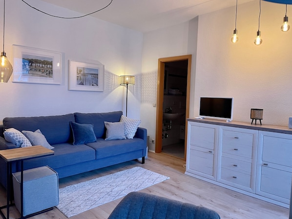 Ein Wohnzimmer mit einer Couch, einem Fernseher, einer Lampe und einem kleinen Schrank.