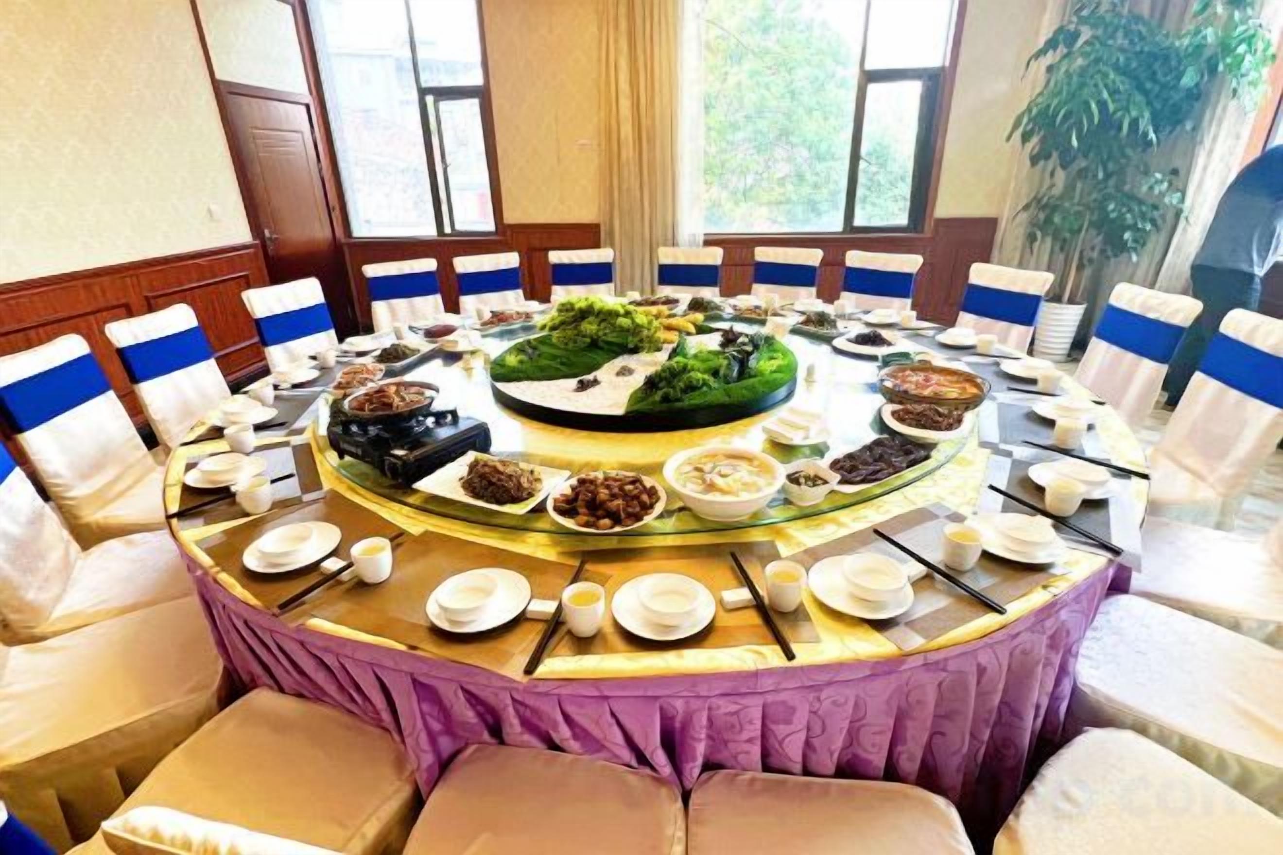 A round table with a central floral arrangement surrounded by various dishes and bowls, set in a room with white chairs and a purple tablecloth.