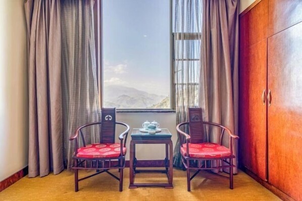 A room with two wooden chairs with red cushions, a small table with a teapot, and a view of mountains through a window with curtains.