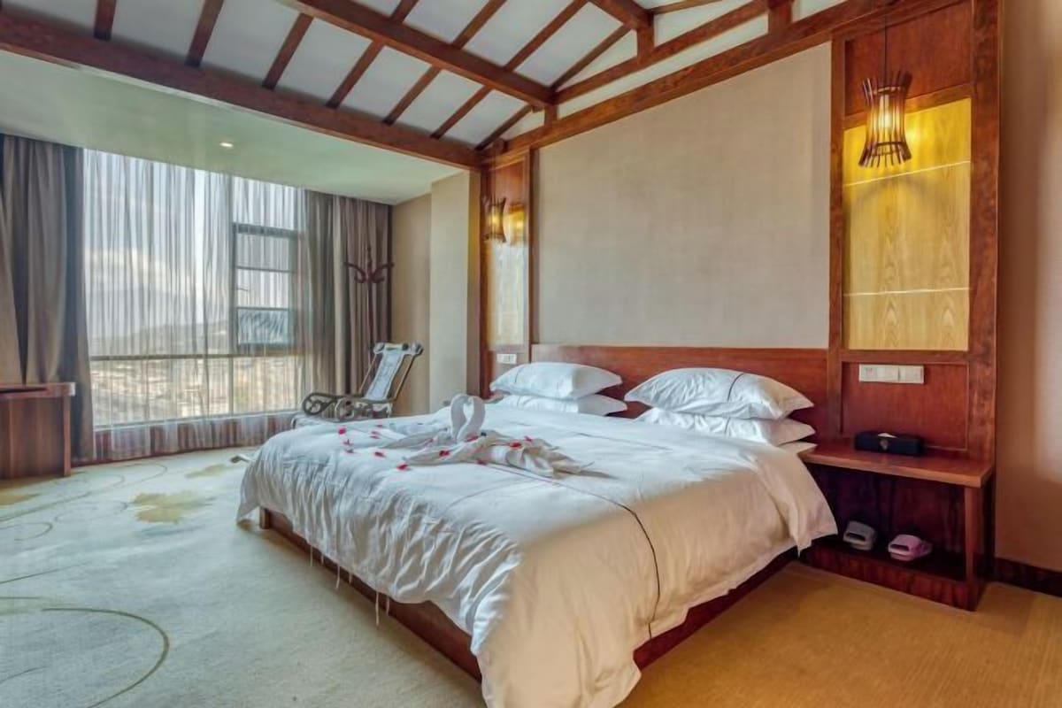 A large bed with white linens and a wooden headboard, a bedside table with a lamp, and a large window with curtains.