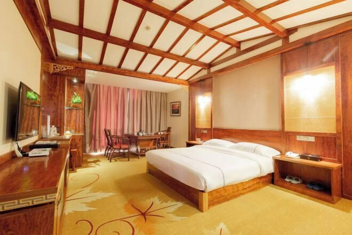 A hotel room with a wooden ceiling, a bed, a desk with a chair, a television, and a bathroom with a mirror and shelf.