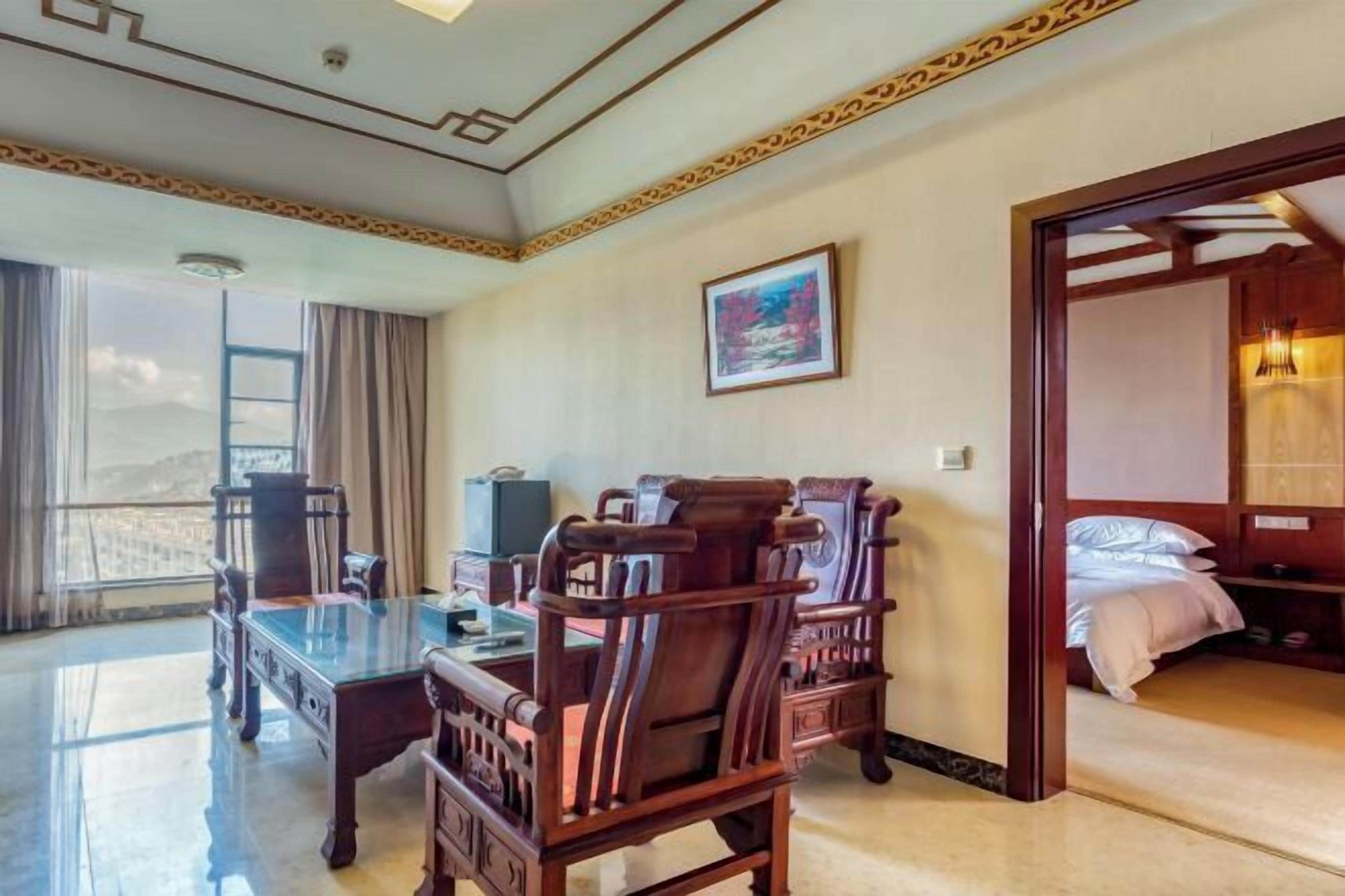 A hotel room with a large window, a wooden desk with chairs, a bed with a wooden headboard, and a painting on the wall.