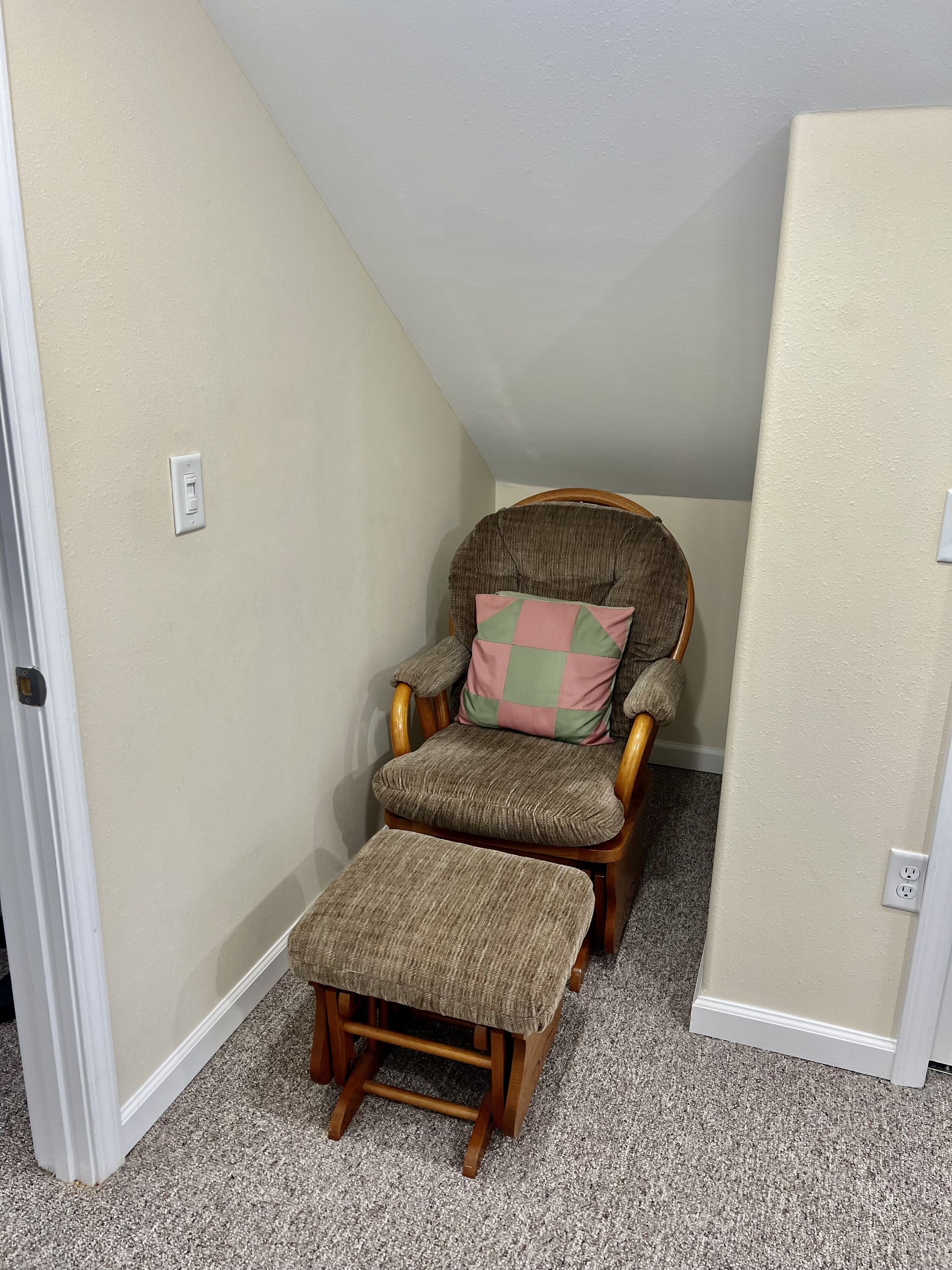 A corner room with a wooden chair and matching ottoman, beige walls, and carpeted floor.
