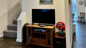 Una sala de estar con una TV de pantalla plana sobre un mueble de madera, un ventilador de techo, una escalera y un pasillo que conduce a otra habitación.