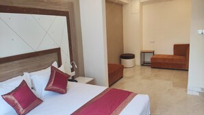 A modern hotel room with a large bed, a wooden headboard, a small table, a chair, and a round ottoman. - HOTEL XENIA (Varanasi)