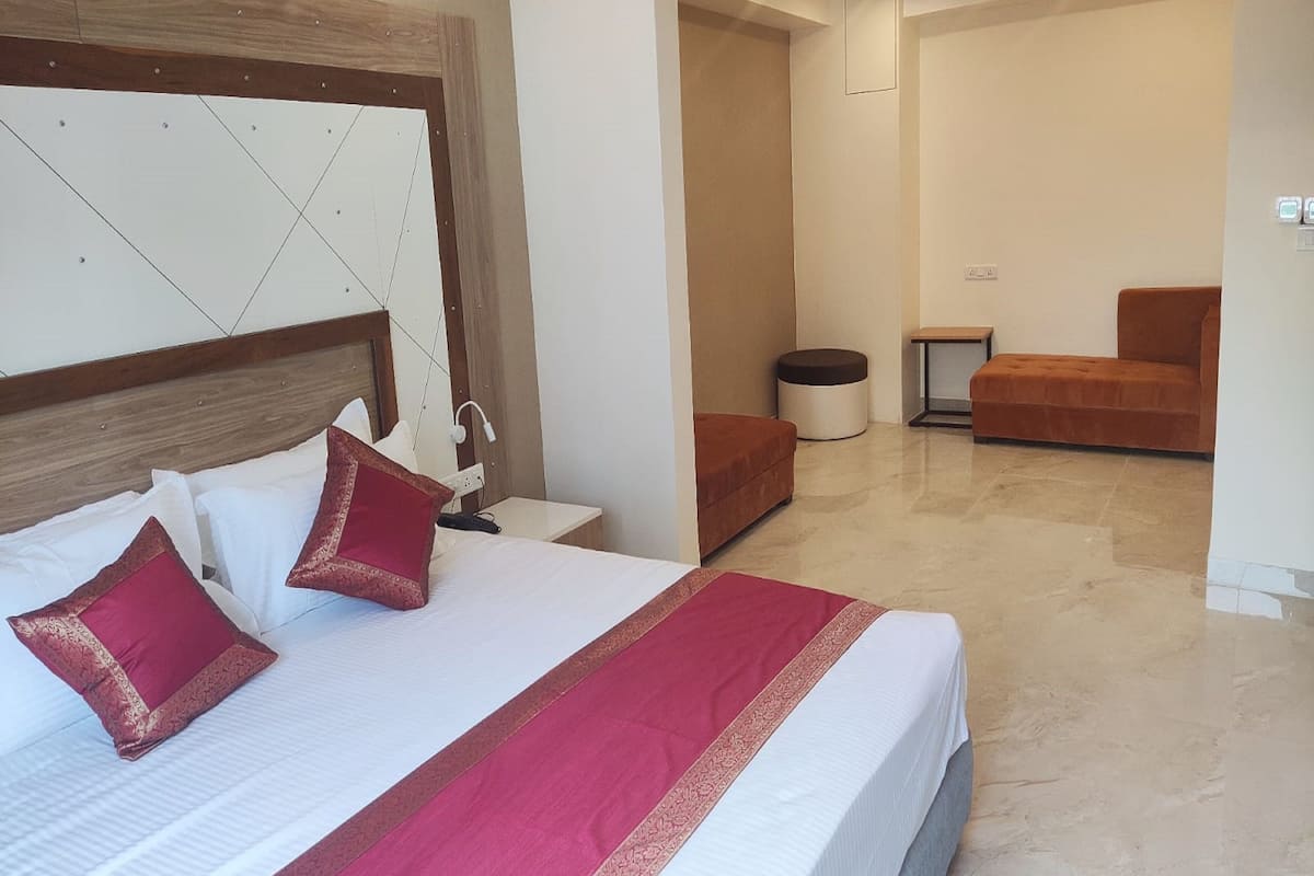 A modern hotel room with a large bed, a wooden headboard, a small table, a chair, and a round ottoman.