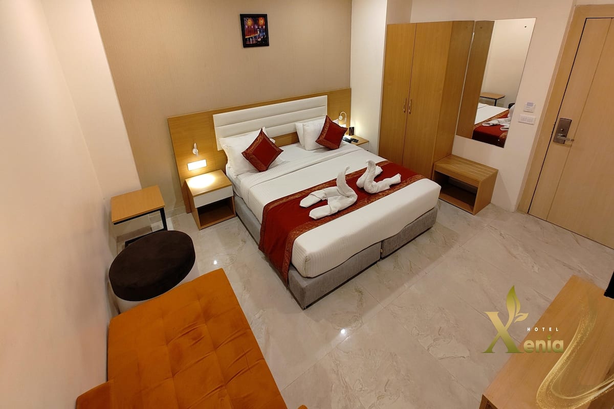 A hotel room with a large bed, a wooden wardrobe, a bedside table, and a small stool.