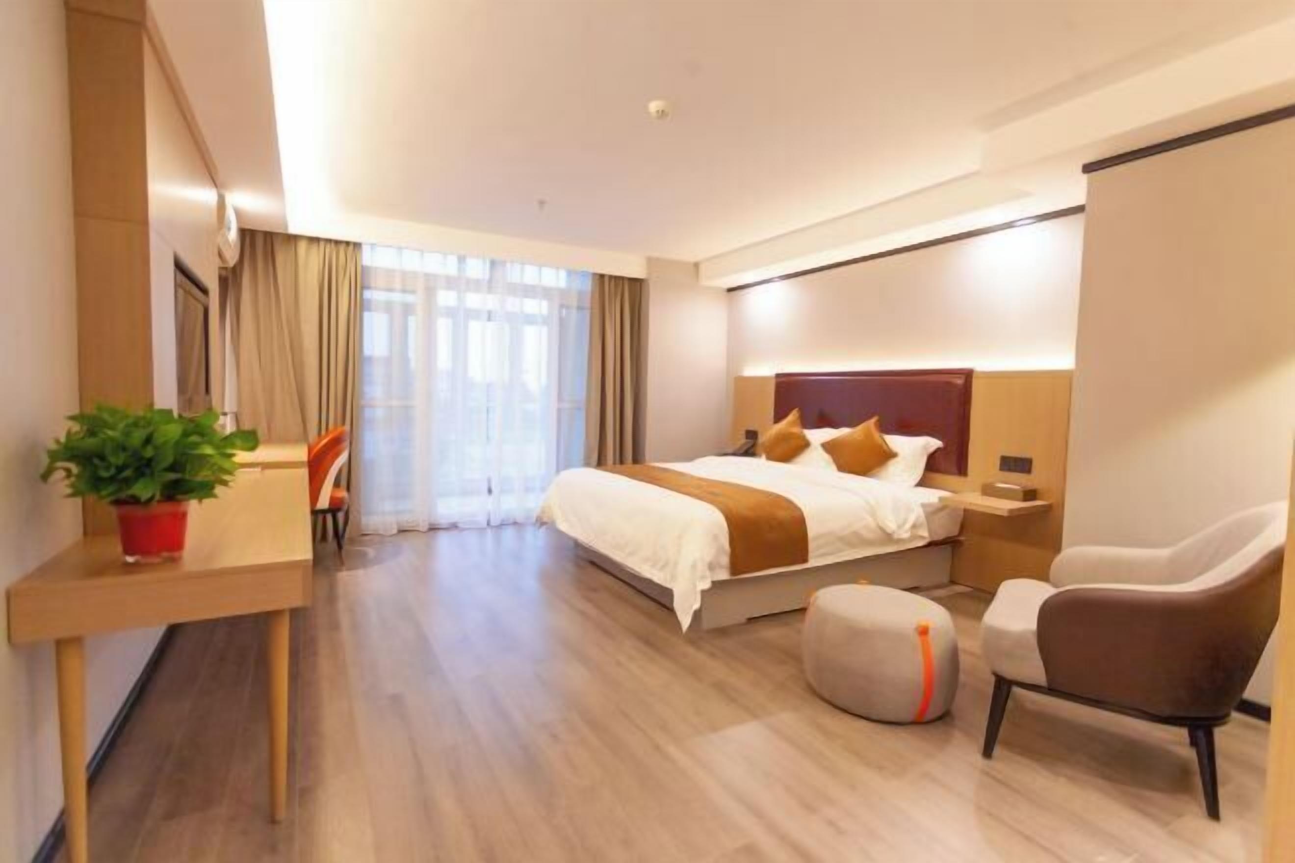 A modern hotel room with a large bed, a desk with a plant, a chair, and a small ottoman.