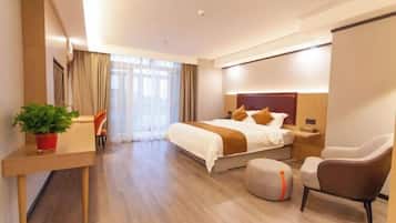A modern hotel room with a large bed, a desk with a plant, a chair, and a small ottoman.