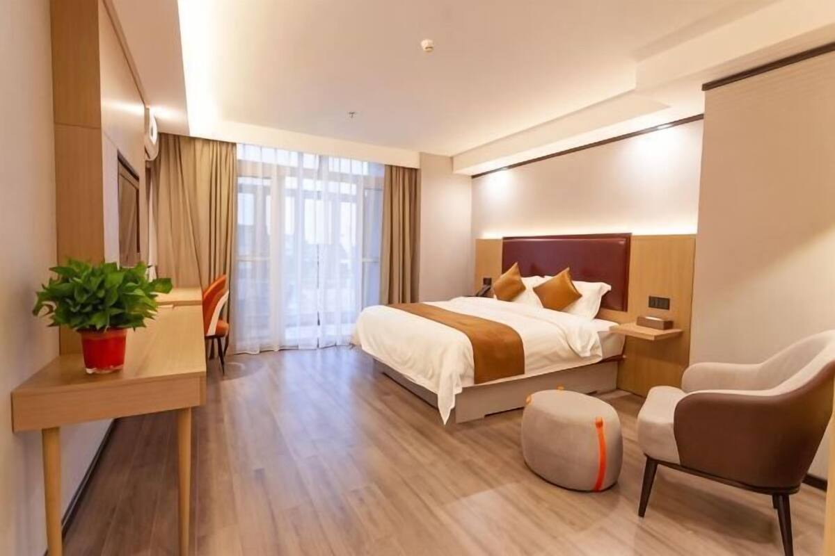 A hotel room with a bed, bedside table, chair, desk, and a potted plant.