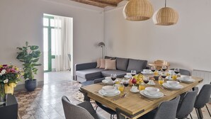 Een eetruimte met een houten tafel gedekt voor een maaltijd, een grijze bank en een groot raam met gordijnen.