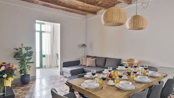 Een eetruimte met een houten tafel gedekt voor een maaltijd, een grijze bank en een groot raam met gordijnen.