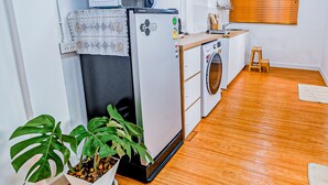 Cocina compacta equipada con refrigerador, microondas y lavadora. Hay un taburete de madera y una planta en maceta.