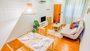 Un comedor con una mesa de madera dispuesta para cuatro personas, un televisor y una planta al fondo.