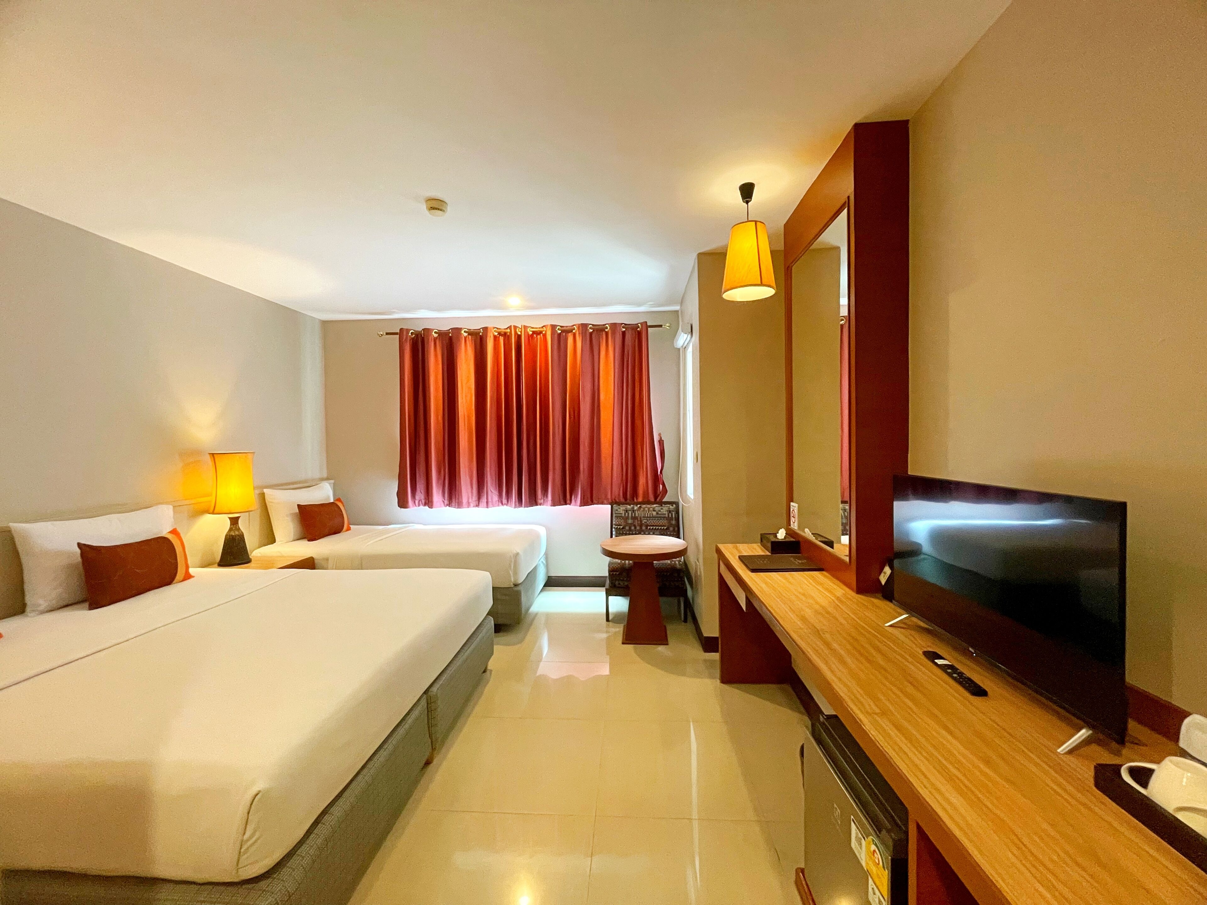 A hotel room with two beds, a desk with a TV, a small table, and a window with red curtains.