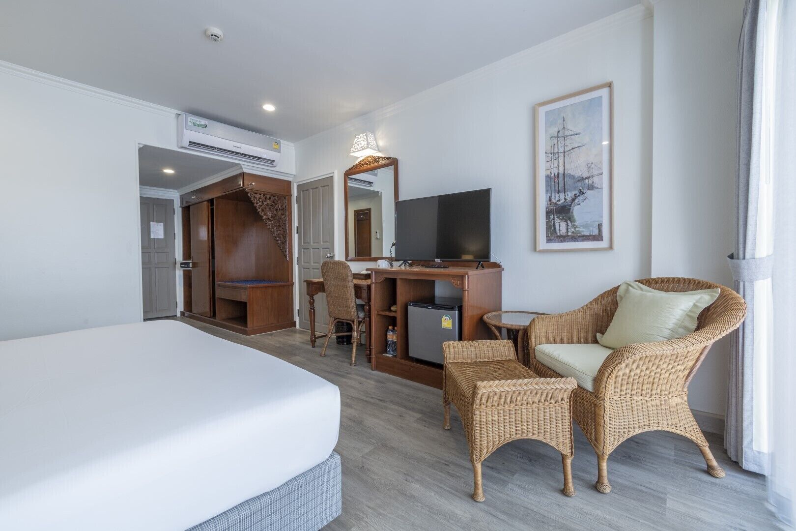 Een hotelkamer met een bed, een bureau met een computer, een televisie, rieten meubels en een schilderij aan de muur.