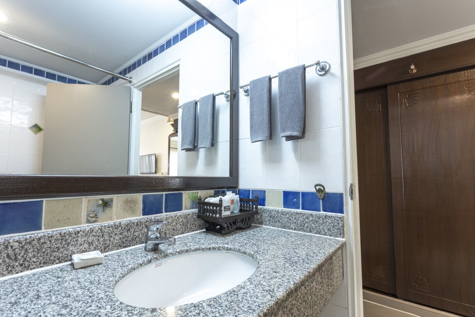 A bathroom with a granite countertop, a white sink, and a mirror. There are towels hanging on a rack and a wooden door.
