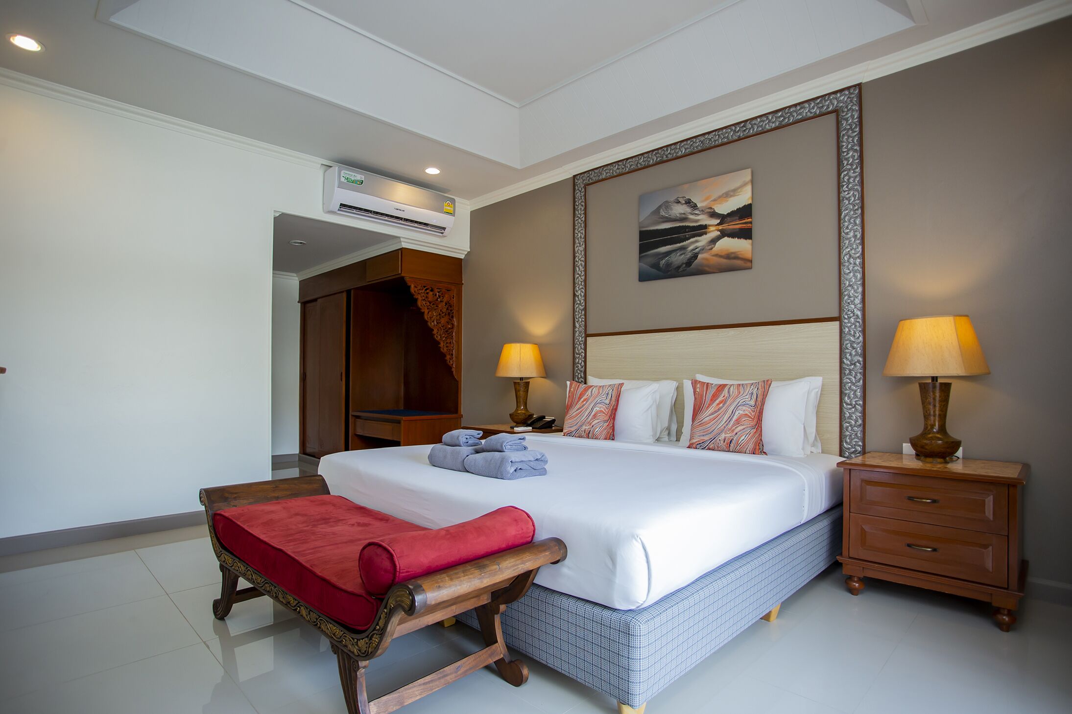 A neatly arranged bedroom with a large bed, bedside tables, a red bench, and an air conditioning unit.