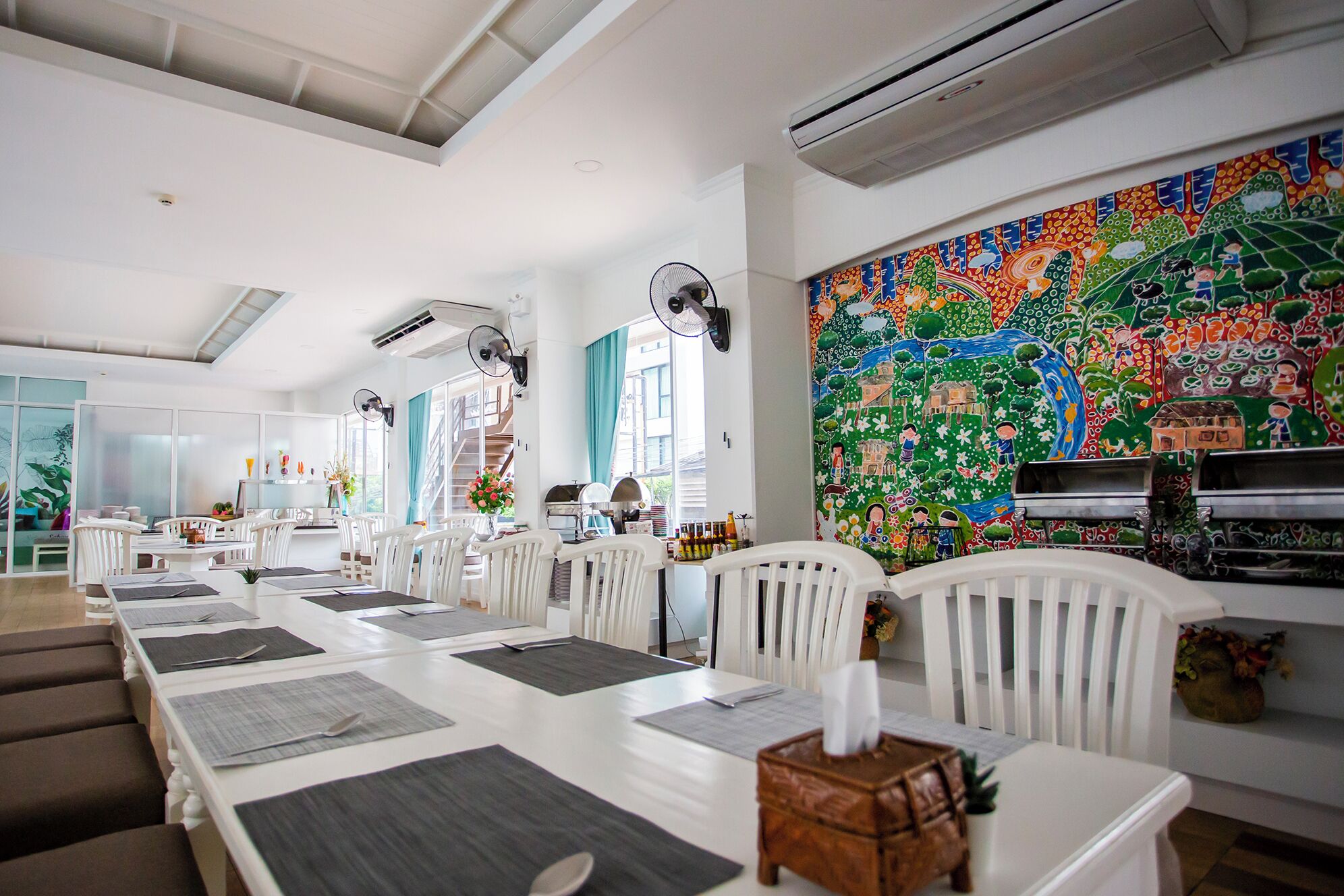 Un espace repas avec des chaises et des tables blanches, des unités de climatisation et une fresque colorée sur le mur.