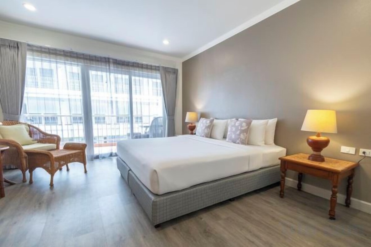 A hotel room with a large bed, a sitting area with a chair, a small table with a lamp, and a view through the window.