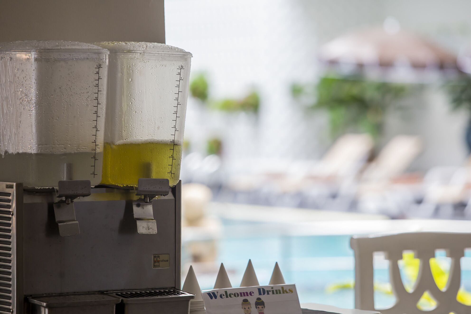 A beverage dispenser with a 'Welcome Drinks' sign, a pool in the background, and a chair with a yellow cushion.