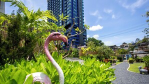 A garden with a pink flamingo statue, lush greenery, and a pathway leading to a building with a blue facade. - Sabai Sabana (Pattaya)