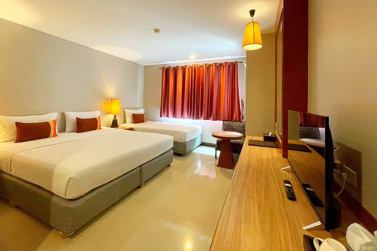 A hotel room with two beds, a desk, a TV, and a window with red curtains.