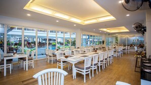 A spacious dining area with white tables and chairs, large windows offering views of the outside, and ceiling fans. - Sabai Sabana (Pattaya)