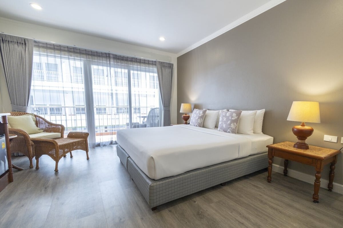 A hotel room with a large bed, a wicker chair, a small table with a lamp, and a view through large windows.