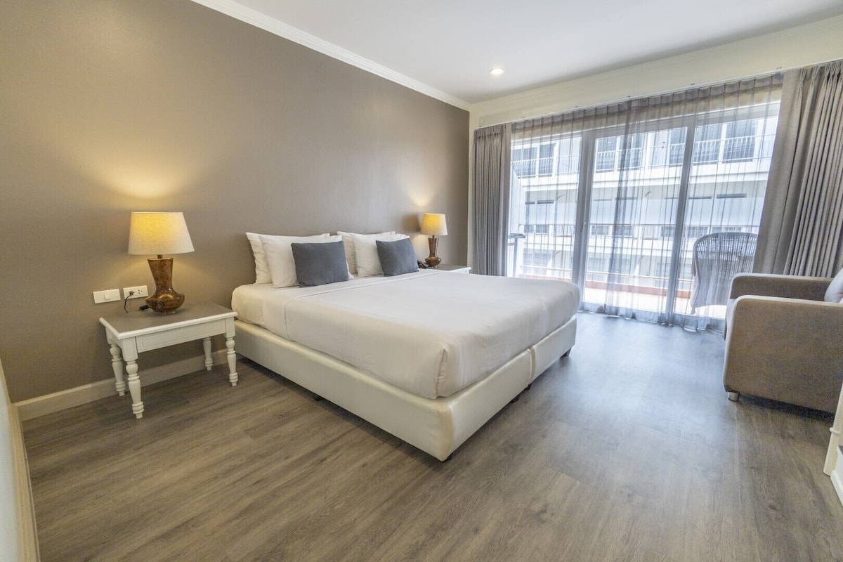 A hotel room with a large bed, two bedside tables with lamps, a chair, and a balcony with curtains.