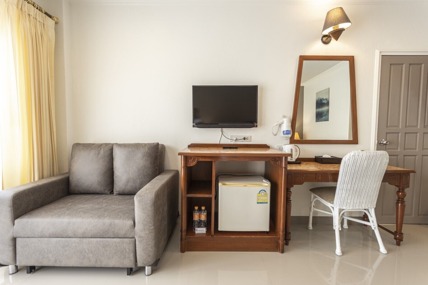 Een hotelkamer met een grijze bank, een houten bureau met een computer en een lamp, een kleine koelkast en een witte stoel.