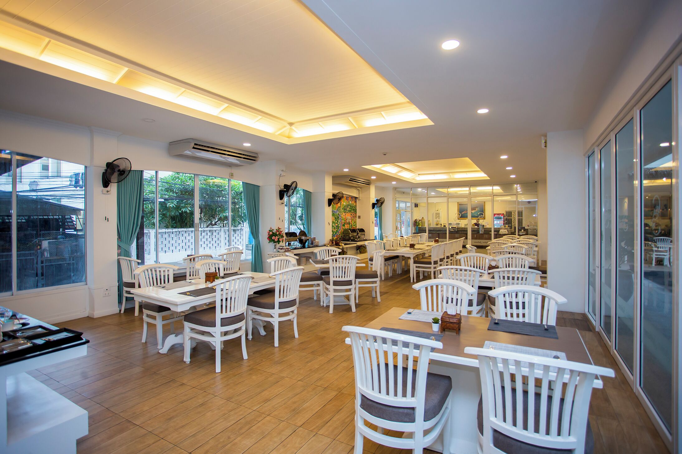 A spacious dining area with multiple tables and white chairs, large windows with curtains, and an air conditioning unit on the wall.