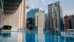 A swimming pool with two people in the water, surrounded by modern high-rise buildings and a tower.