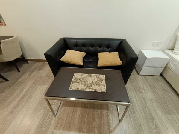 A black leather sofa with two beige cushions, a small table with a lace table runner, and a white bedside table with a lamp.