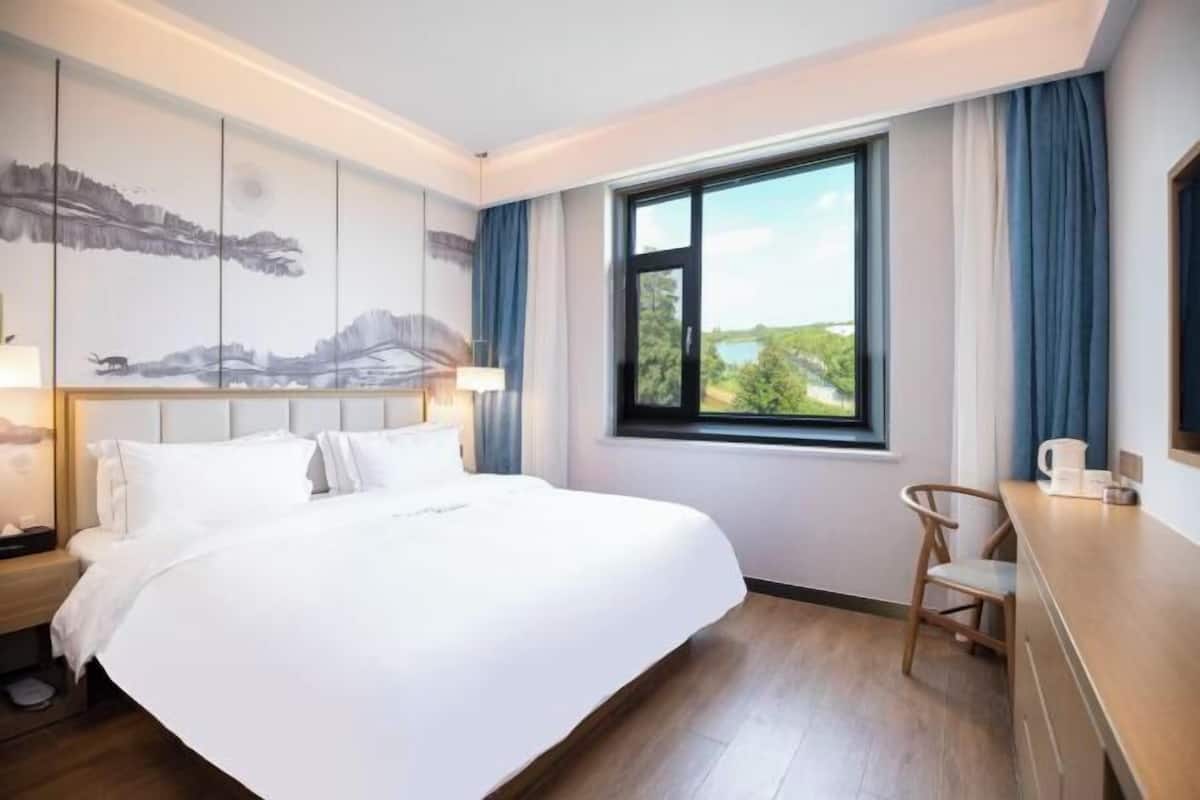 A hotel room with a large bed, a desk, a chair, a window with a view, and a painting on the wall.