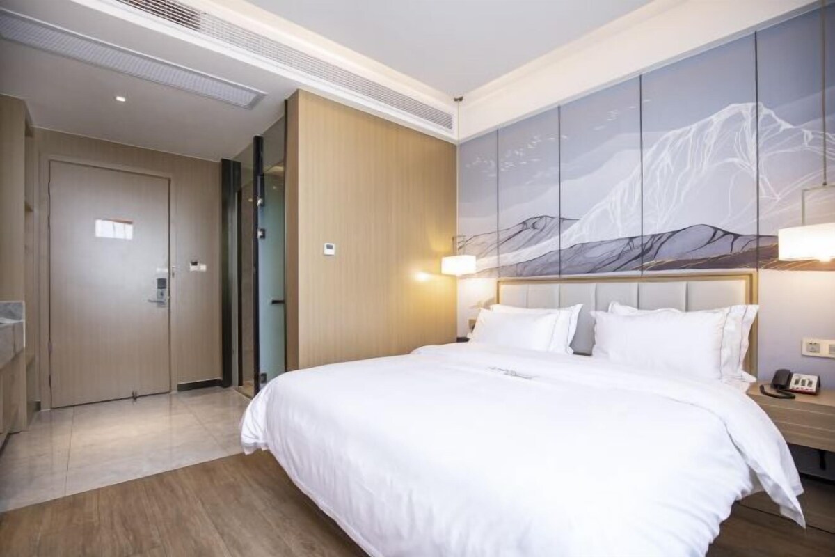 A hotel room with a large bed, a wooden wardrobe, a wall-mounted artwork, and a bedside table with a lamp.
