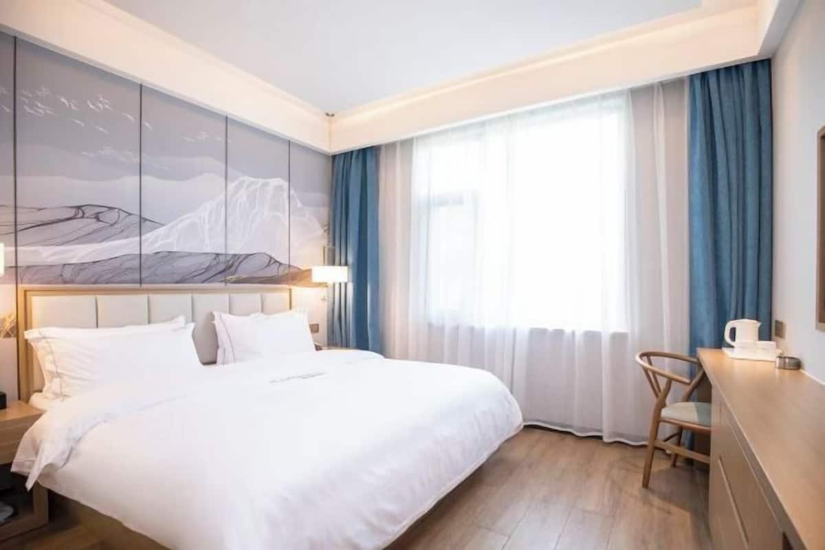 A hotel room with a bed, a desk, a chair, a nightstand, and a wall with a mountain painting.