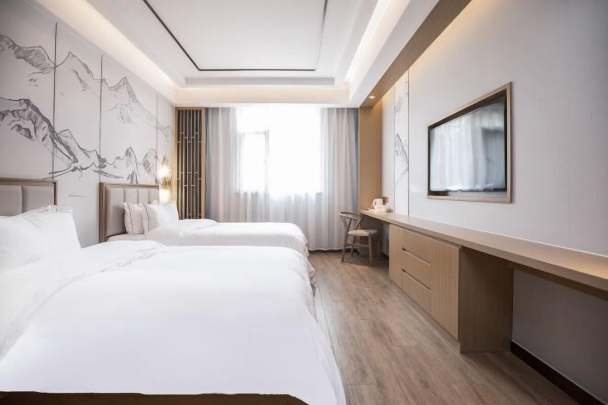 A hotel room with two beds, a desk, a chair, a TV, and a wall with a mountain painting.