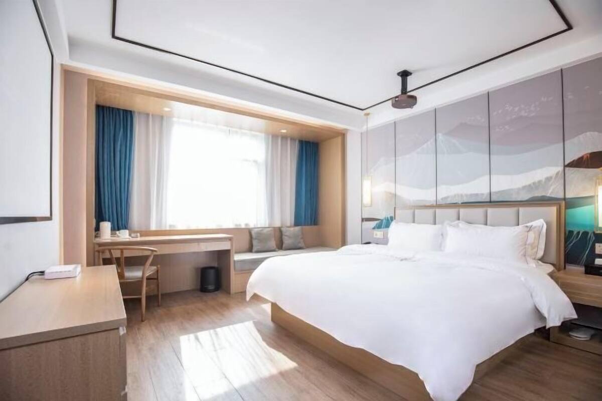 A modern hotel room with a large bed, a desk with a chair, a window with curtains, and a wall with a mountain view.