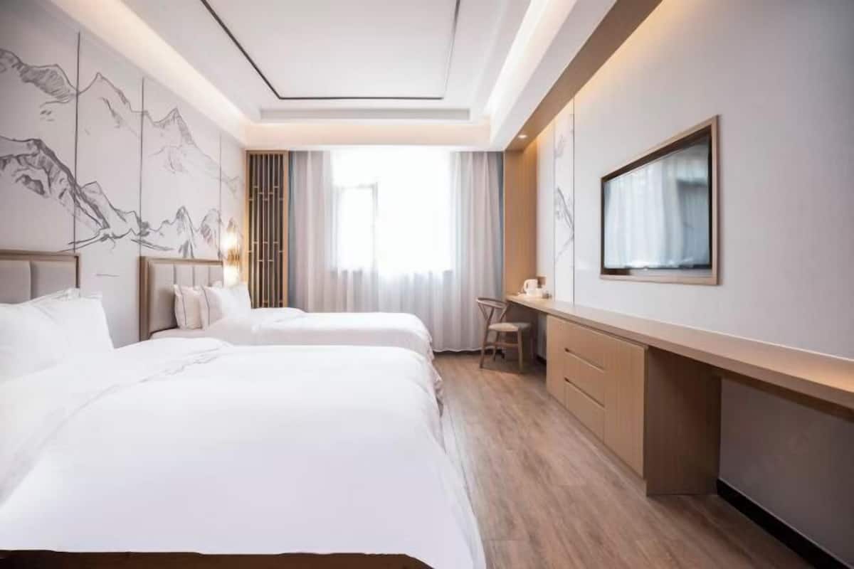 A hotel room with two beds, a desk, a chair, a TV, and a large window with curtains.