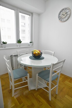 A round glass table with four white chairs, a fruit bowl, and two potted plants on a white window sill. - Modern & Cozy near City Center ~ Balcony ~ Parking (Skopje)