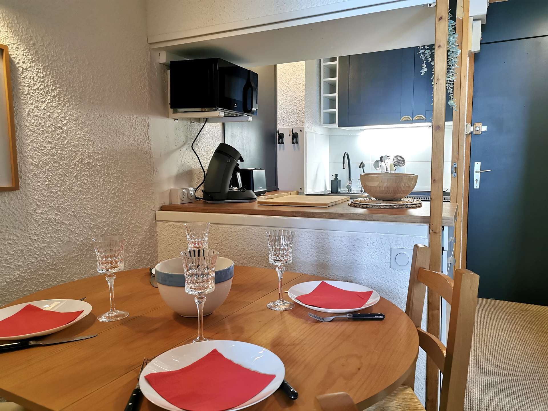 A dining area with a wooden table set for four, including wine glasses and plates. A kitchen is visible in the background with a microwave and cabinets.