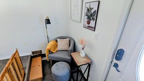 Ett hörnrum med en grå fåtölj, en liten pall, en golvlampa, en trästol, ett litet bord med en vas och en dörr med ett blått handtag.