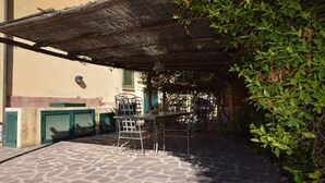 En uteplats med stengolv, ett metallbord och stolar, en pergola som ger skugga och en vägg med ett fönster och en lampa.