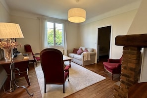 A living room with a wooden floor, a dining table with chairs, a sofa, a fireplace, and a large window.